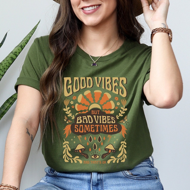 May include: Green t-shirt with a retro graphic design that says "Good Vibes But Bad Vibes Sometimes And That's Ok". The graphic features a sun, flowers, mushrooms, and lightning bolts.
