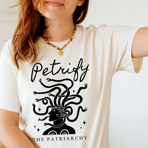 K&ouml;nnte beinhalten: Wei&szlig;es T-Shirt mit einer schwarzen Grafik von Medusa mit Schlangen als Haaren. Der Text "Petrify the Patriarchy" ist unter der Grafik gedruckt.