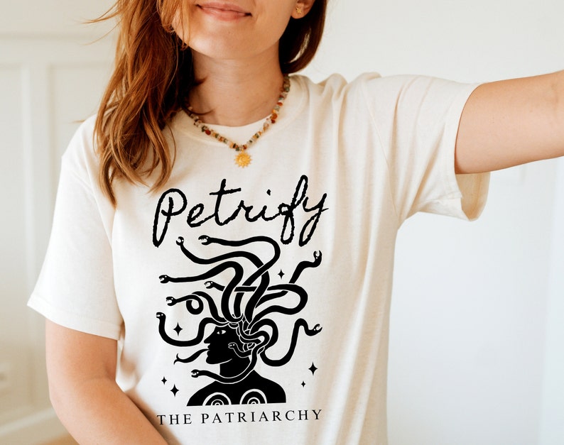 K&ouml;nnte beinhalten: Wei&szlig;es T-Shirt mit einer schwarzen Grafik von Medusa mit Schlangen als Haaren. Der Text "Petrify the Patriarchy" ist unter der Grafik gedruckt.