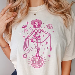 May include: Cream-colored t-shirt featuring a pink graphic of a cowgirl in a space-themed design. The cowgirl is holding a lasso and standing on a disco ball. The design includes a UFO, planets, and stars.