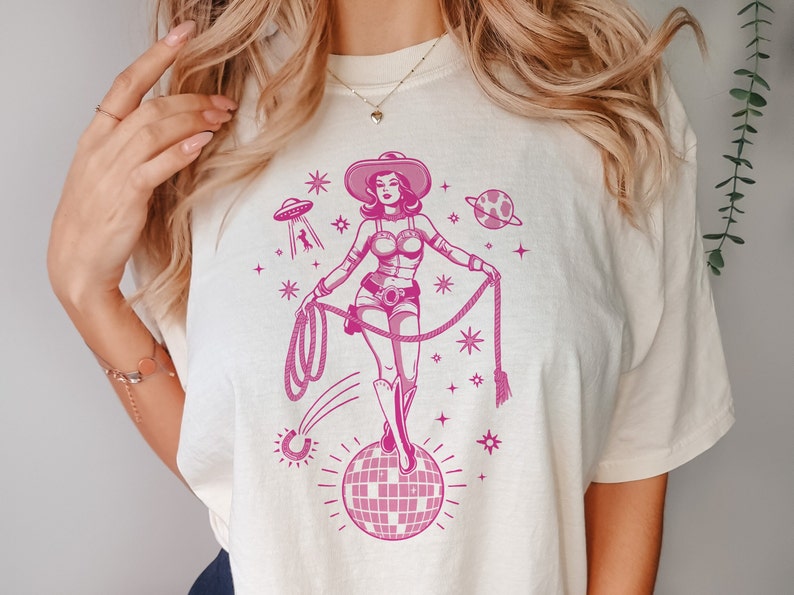 May include: Cream-colored t-shirt featuring a pink graphic of a cowgirl in a space-themed design. The cowgirl is holding a lasso and standing on a disco ball. The design includes a UFO, planets, and stars.