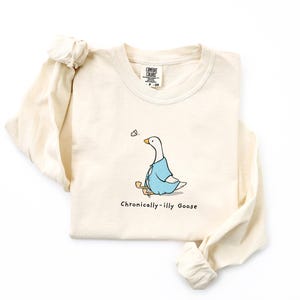 May include: A beige long-sleeved t-shirt with a cartoon illustration of a goose wearing a blue shirt and holding a stick. The text "Chronically-illy Goose" is printed below the goose.