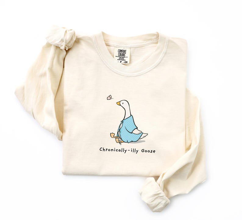 May include: A beige long-sleeved t-shirt with a cartoon illustration of a goose wearing a blue shirt and holding a stick. The text "Chronically-illy Goose" is printed below the goose.