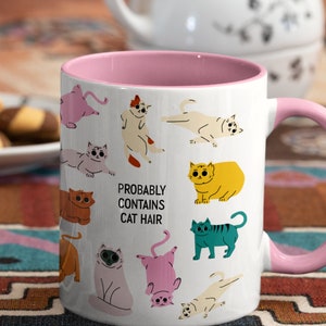 May include: White ceramic mug with a pink rim and handle. The mug features a whimsical design of colourful cats and the text "Probably Contains Cat Hair".