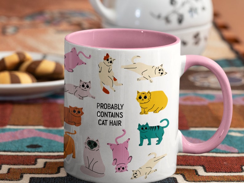 May include: White ceramic mug with a pink rim and handle. The mug features a whimsical design of colourful cats and the text "Probably Contains Cat Hair".