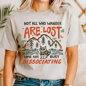 May include: A light grey t-shirt with a graphic design featuring a raccoon in a forest setting. The text on the shirt reads "Not all who wander *are* lost. Some are busy dissociating."