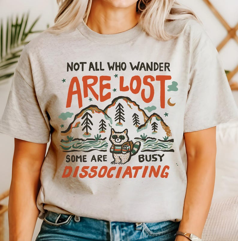 May include: A light grey t-shirt with a graphic design featuring a raccoon in a forest setting. The text on the shirt reads "Not all who wander *are* lost. Some are busy dissociating."