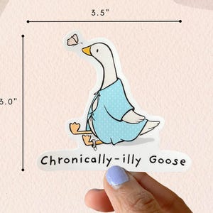 May include: A white goose wearing a light blue polka dot robe with a butterfly on its wing. The goose is sitting down with its legs crossed. The text "Chronically-illy Goose" is written below the goose. The sticker is 3 inches wide and 3.5 inches tall.