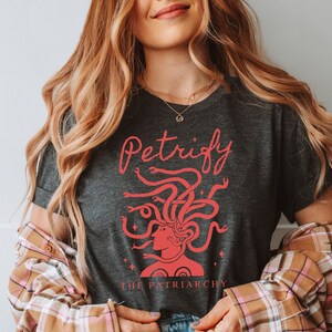 K&ouml;nnte beinhalten: Ein dunkelgraues T-Shirt mit einer roten Grafik von Medusas Kopf mit Schlangen als Haaren. Der Text "Petrify the Patriarchy" ist in rot unter der Grafik gedruckt.