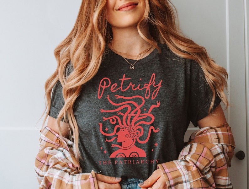 K&ouml;nnte beinhalten: Ein dunkelgraues T-Shirt mit einer roten Grafik von Medusas Kopf mit Schlangen als Haaren. Der Text "Petrify the Patriarchy" ist in rot unter der Grafik gedruckt.