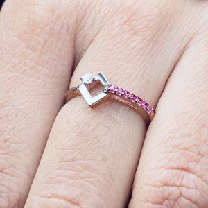 Op de afbeelding: Een roségouden ring met een hartvormig ontwerp en een kleine diamant. De ring heeft ook een rij kleine roze edelstenen. De ring zit om een vinger.