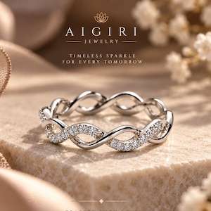 Puede incluir: Un anillo plateado con diseño de infinito entrelazado y una serie de pequeñas piedras claras y brillantes a lo largo de la banda. El texto AIGIRI JEWELRY, TIMELESS SPARKLE FOR EVERY TOMORROW y CRAFTED WITH LOVE MADE TO LAST es visible.
