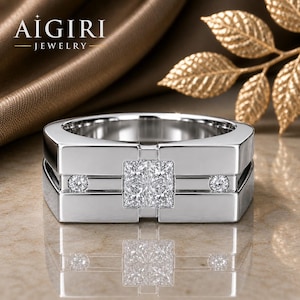 May include: A silver ring featuring a square-cut diamond at the centre and two smaller round diamonds. The ring has a contemporary, geometric design. The brand name "AIGIRI - JEWELRY -" is visible in the upper left corner.