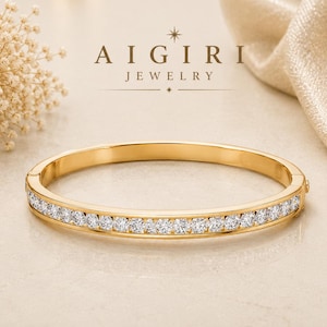 Puede incluir: Una pulsera rígida articulada de tono dorado con una hilera de piedras claras de corte redondo engastadas en la parte frontal. El texto AIGIRI JEWELRY aparece sobre la pulsera en fuente oscura, junto a pequeños detalles gráficos de estrellas.