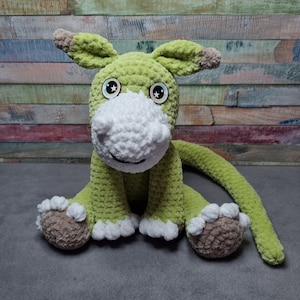 May include: A green crocheted stuffed animal with white accents and large eyes. The animal has a long tail and is sitting on a gray surface.