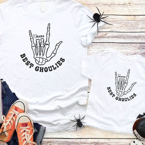 May include: Two white t-shirts with a skeleton hand making the "I love you" sign language gesture. The shirts say "Best Ghoulies" and have the names "Mama" and "Harper" in a heart shape on the hand. The shirts are on a wooden surface with orange sneakers, blue jeans, and black spiders.
