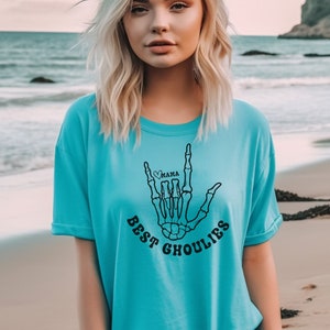 May include: A turquoise blue t-shirt with a black skeleton hand graphic. The hand is making the "I love you" sign language with a small heart and the word "mama" inside. The text "Best Ghoulies" is printed below the hand.