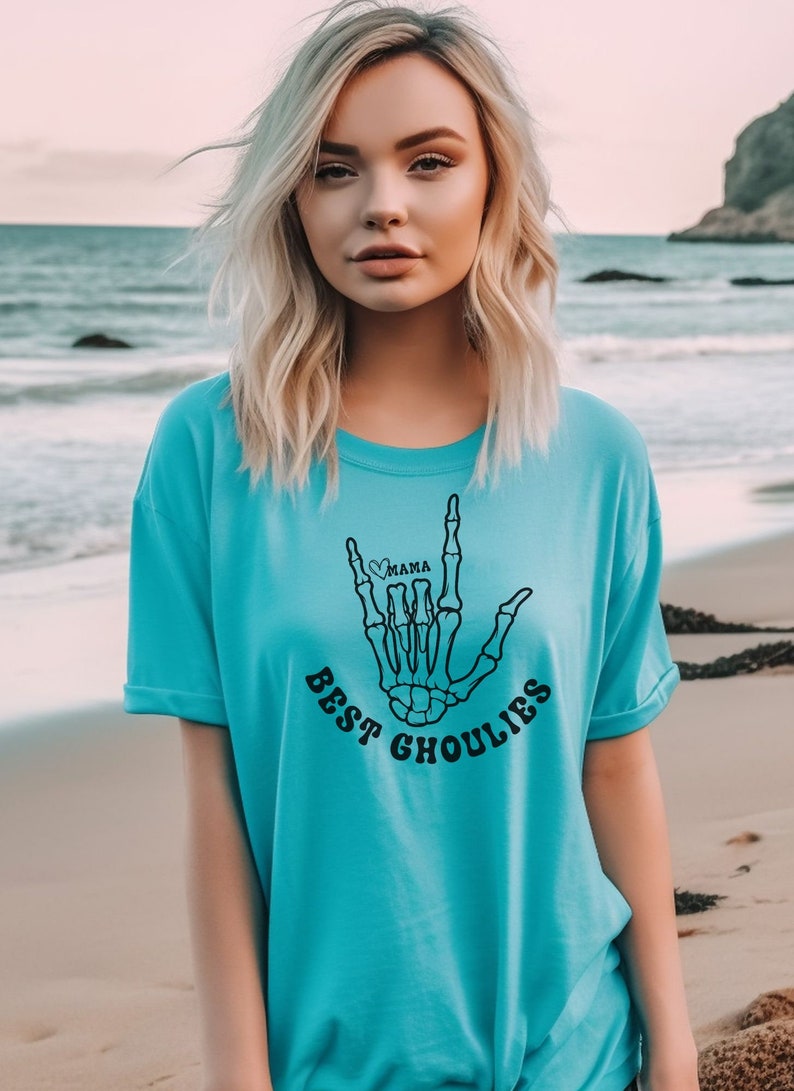 May include: A turquoise blue t-shirt with a black skeleton hand graphic. The hand is making the "I love you" sign language with a small heart and the word "mama" inside. The text "Best Ghoulies" is printed below the hand.