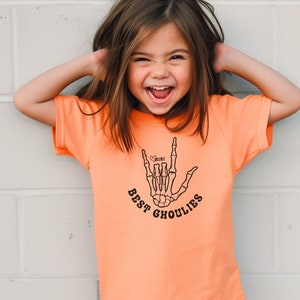 May include: A young girl wearing an orange t-shirt with a black skeleton hand graphic and the text "Best Ghoulies" and a small heart with the word "mini" inside.