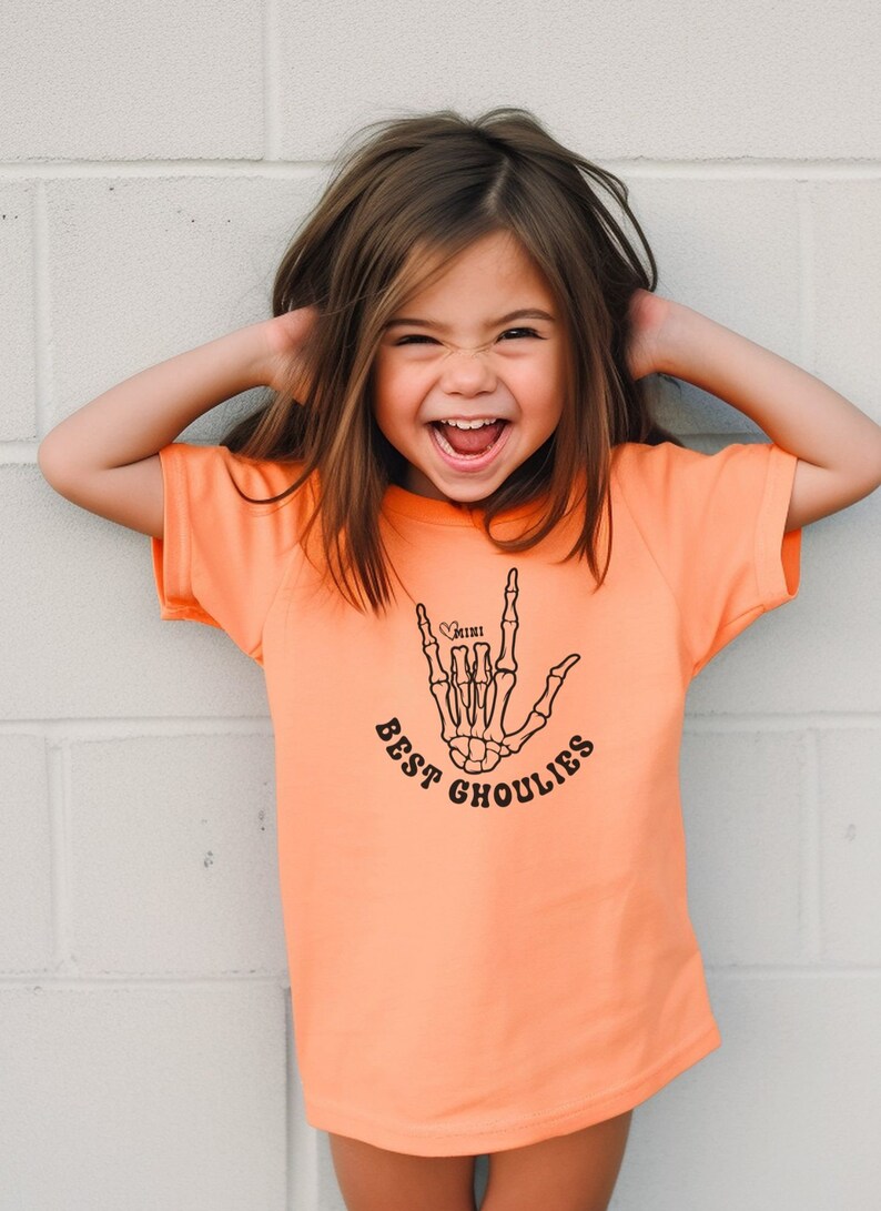 May include: A young girl wearing an orange t-shirt with a black skeleton hand graphic and the text "Best Ghoulies" and a small heart with the word "mini" inside.