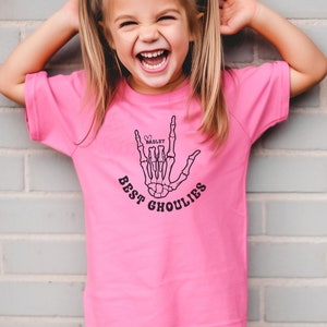 May include: A pink t-shirt with a black skeleton hand making the "I love you" sign. The hand has a heart with the name "HADLEY" inside. The text "BEST GHOULIES" is written below the hand.