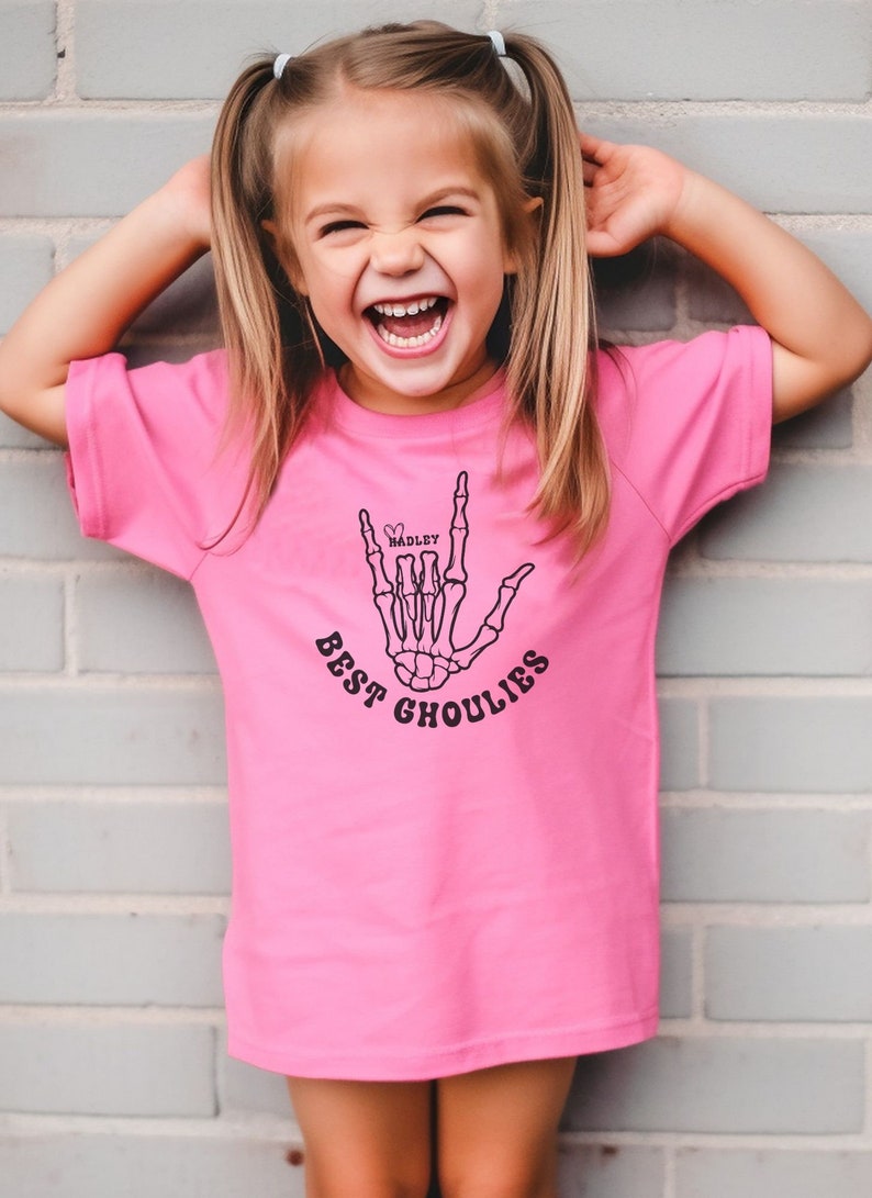 May include: A pink t-shirt with a black skeleton hand making the "I love you" sign. The hand has a heart with the name "HADLEY" inside. The text "BEST GHOULIES" is written below the hand.