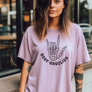 May include: A light purple t-shirt with a skeleton hand making the "I love you" sign in American Sign Language. The hand has a heart with the word "mama" inside. The text "Best Ghoulies" is printed below the hand.