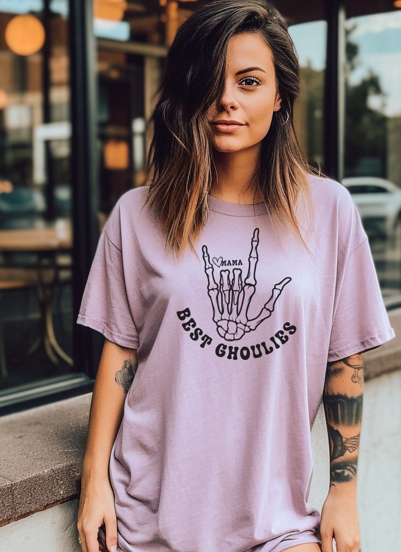May include: A light purple t-shirt with a skeleton hand making the "I love you" sign in American Sign Language. The hand has a heart with the word "mama" inside. The text "Best Ghoulies" is printed below the hand.