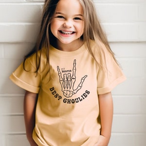 May include: A young girl wearing a light yellow t-shirt with a black skeleton hand graphic and the text "BEST GHOULIES" and the name "HARPER" in a heart.