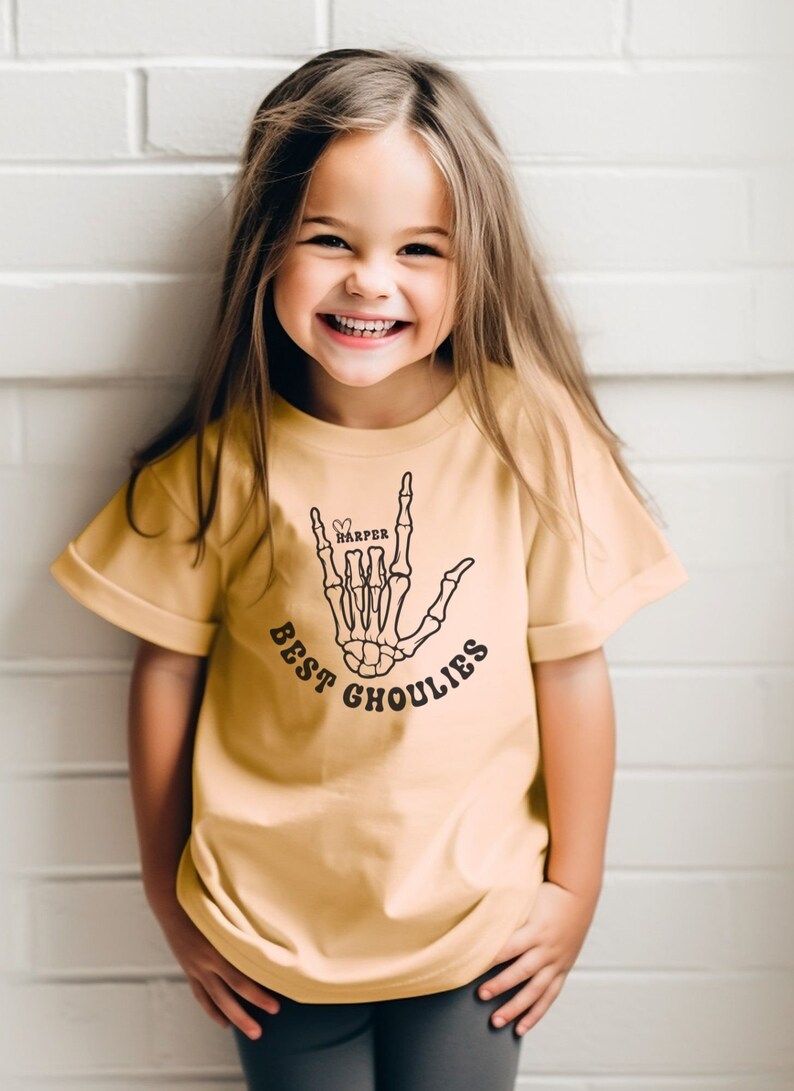 May include: A young girl wearing a light yellow t-shirt with a black skeleton hand graphic and the text "BEST GHOULIES" and the name "HARPER" in a heart.