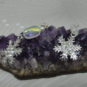May include: A pair of silver snowflake earrings with iridescent glass beads. The earrings are displayed on a bed of amethyst crystals.