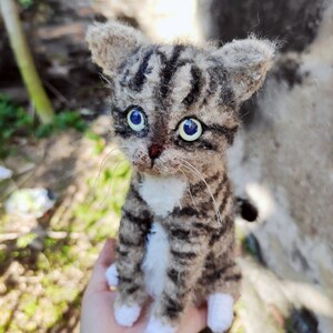 May include: A small, crocheted tabby cat with white paws and a white chest. The cat has blue eyes and is sitting on a person's hand.
