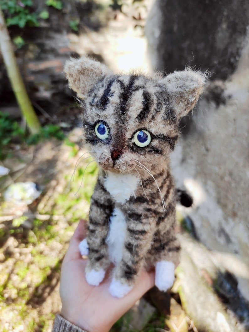 May include: A small, crocheted tabby cat with white paws and a white chest. The cat has blue eyes and is sitting on a person's hand.