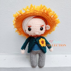 Könnte beinhalten: Eine gehäkelte Puppe mit einem gelben Hut, einer blauen Jacke, grauen Hosen und einer Sonnenblumennadel. Die Puppe hat einen roten Bart und einen weißen Verband auf dem Kopf. Die Puppe sitzt auf einer weißen Oberfläche. PJ Crochet Collection.