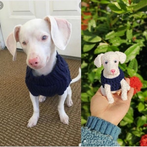 May include: A white dog wearing a blue knitted sweater. The dog is sitting on a brown carpet. A crocheted white dog wearing a blue knitted sweater is being held in a hand.