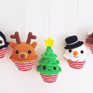 May include: Five crocheted Christmas decorations shaped like cupcakes with different festive characters on top. The decorations include a penguin, reindeer, Christmas tree, snowman, and a gingerbread man.