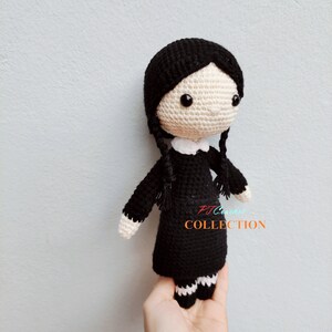 Pu&ograve; includere: Una bambola di uncinetto nera con dettagli bianchi. La bambola ha i capelli neri, un colletto bianco e gambe a righe bianche e nere. La bambola tiene un cartello che dice "PJ Crochet Collection".