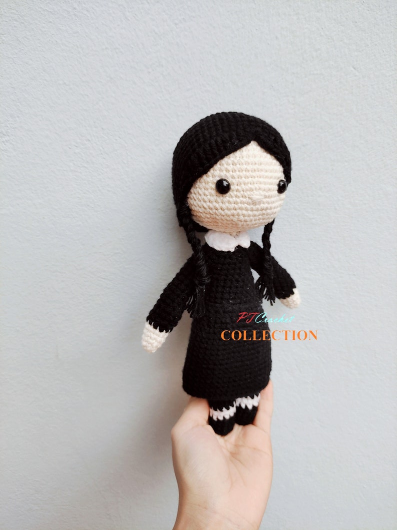 Pu&ograve; includere: Una bambola di uncinetto nera con dettagli bianchi. La bambola ha i capelli neri, un colletto bianco e gambe a righe bianche e nere. La bambola tiene un cartello che dice "PJ Crochet Collection".