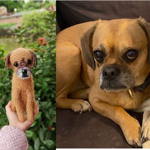 Op de afbeelding: Een gehaakt hondenspeelgoed, bruin en beige van kleur, wordt in de hand van een persoon gehouden. Het speelgoed is een realistische weergave van een hond, met een bruine neus en zwarte ogen. De achtergrond is een wazig beeld van groen gebladerte.