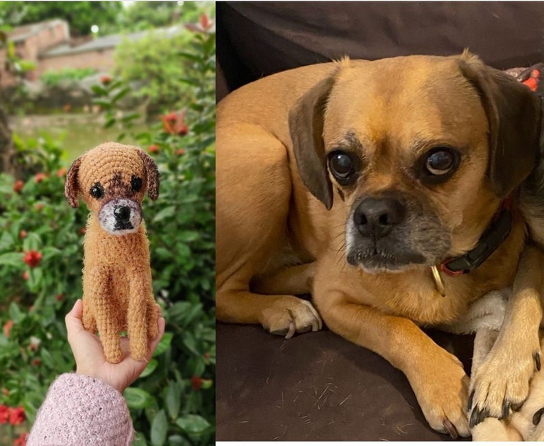 Op de afbeelding: Een gehaakt hondenspeelgoed, bruin en beige van kleur, wordt in de hand van een persoon gehouden. Het speelgoed is een realistische weergave van een hond, met een bruine neus en zwarte ogen. De achtergrond is een wazig beeld van groen gebladerte.