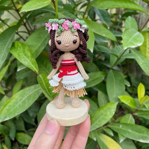 May include: A crocheted doll with brown hair, wearing a red and white dress and a flower crown. The doll is standing on a wooden base.