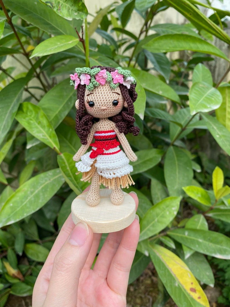May include: A crocheted doll with brown hair, wearing a red and white dress and a flower crown. The doll is standing on a wooden base.