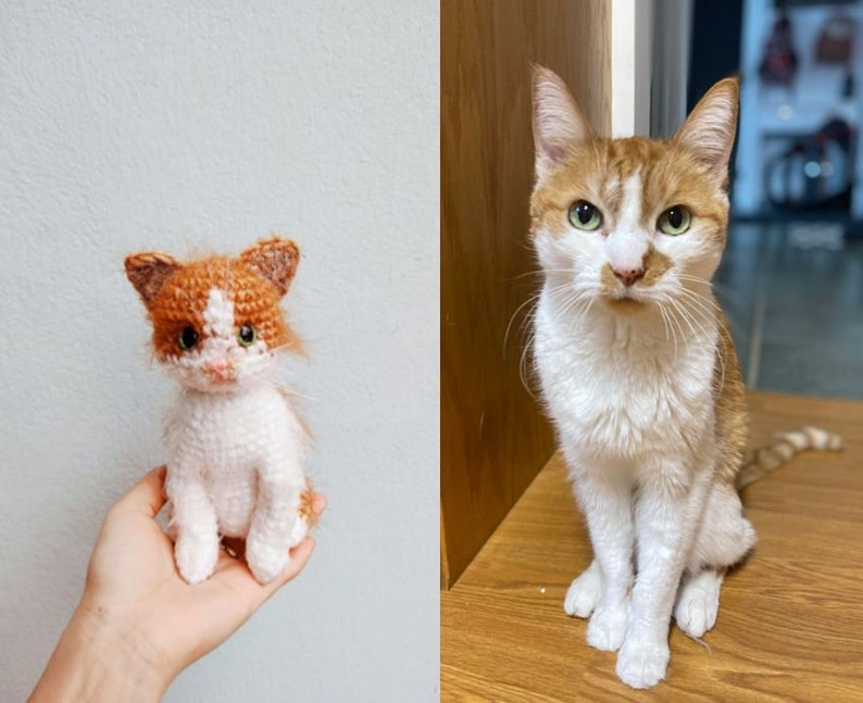 May include: A crocheted orange and white cat toy sitting in a hand. Next to it is a real orange and white cat sitting on a wooden surface.