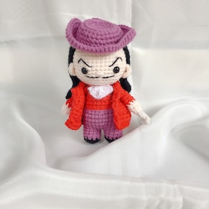 Könnte beinhalten: Eine gehäkelte Amigurumi-Puppe mit einem lila Hut, einer roten Jacke und einer lila Hose. Die Puppe hat einen schelmischen Ausdruck mit schwarzen Augen und einem Schnurrbart.