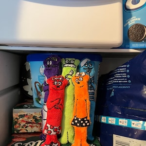 May include: A refrigerator interior with several colorful, plush toy figures standing in front of containers of ice cream and frozen food. The figures are various colors, including red, orange, green, purple, and blue, and are wearing small skirts. The background includes ice cream containers and frozen food boxes.