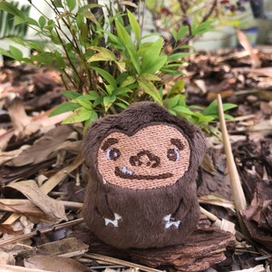 May include: A brown plush toy in the shape of a cartoon Bigfoot, with embroidered facial features and small white claws. The Bigfoot is sitting on a piece of wood, with green foliage in the background.