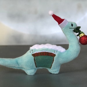 May include: A plush, light blue dinosaur toy with a red and white Santa hat. The dinosaur holds a red Christmas ornament and greenery in its mouth. The dinosaur has a green and brown patch on its back with the word "ICE CREAM".
