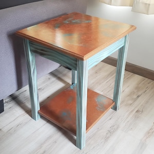 Pode incluir: Uma mesa lateral quadrada com um tampo cor de cobre e uma prateleira inferior. A estrutura da mesa é pintada em um tom verde-azulado claro, com um acabamento de cobre manchado no tampo e na prateleira. A mesa tem aproximadamente 51 cm de altura.