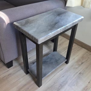 May include: A small, rectangular side table with a gray marble-look top and a dark brown wooden frame. The table has a lower shelf and is positioned next to a gray sofa. The table's dimensions are approximately 20 inches long, 12 inches wide, and 24 inches high.