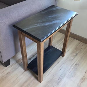 May include: A small, rectangular side table with a dark gray top and a lower shelf. The table has a wooden frame with a natural finish. The top and shelf appear to be made of a dark, possibly stone-like material. The table is approximately 24 inches tall.
